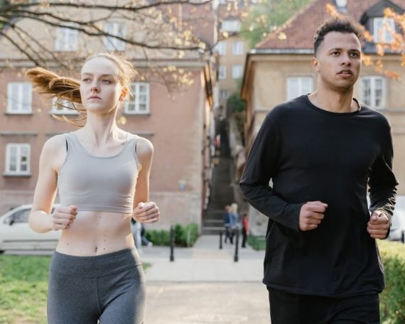 A young woman and man jogging on a city street during the day, wearing sportswear.